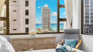A hotel room with a bed, a chair, a dresser, and a view of a tall building and the ocean.