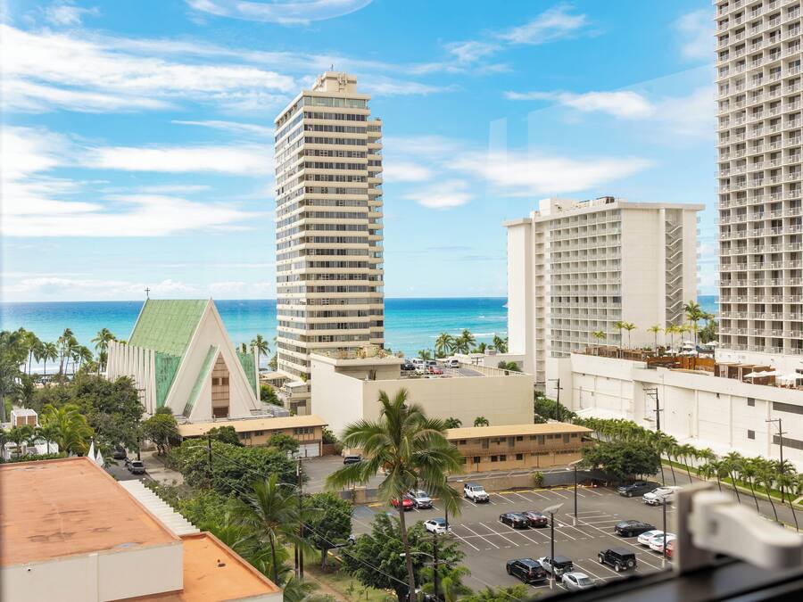 A view of a hotel with a parking lot, a tall building, and a beach in the background.