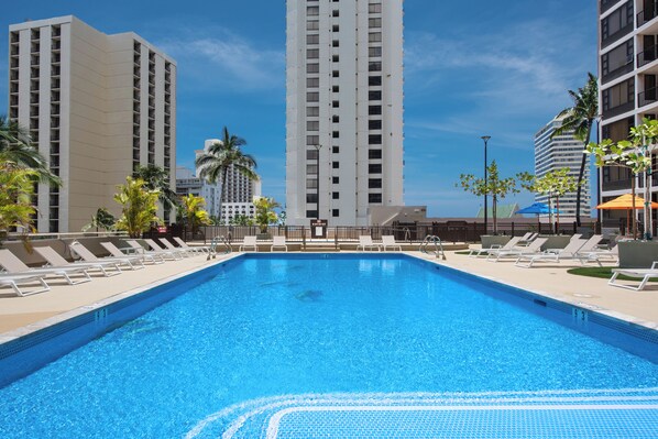 Piscina com espreguiçadeiras e edifícios altos ao fundo.