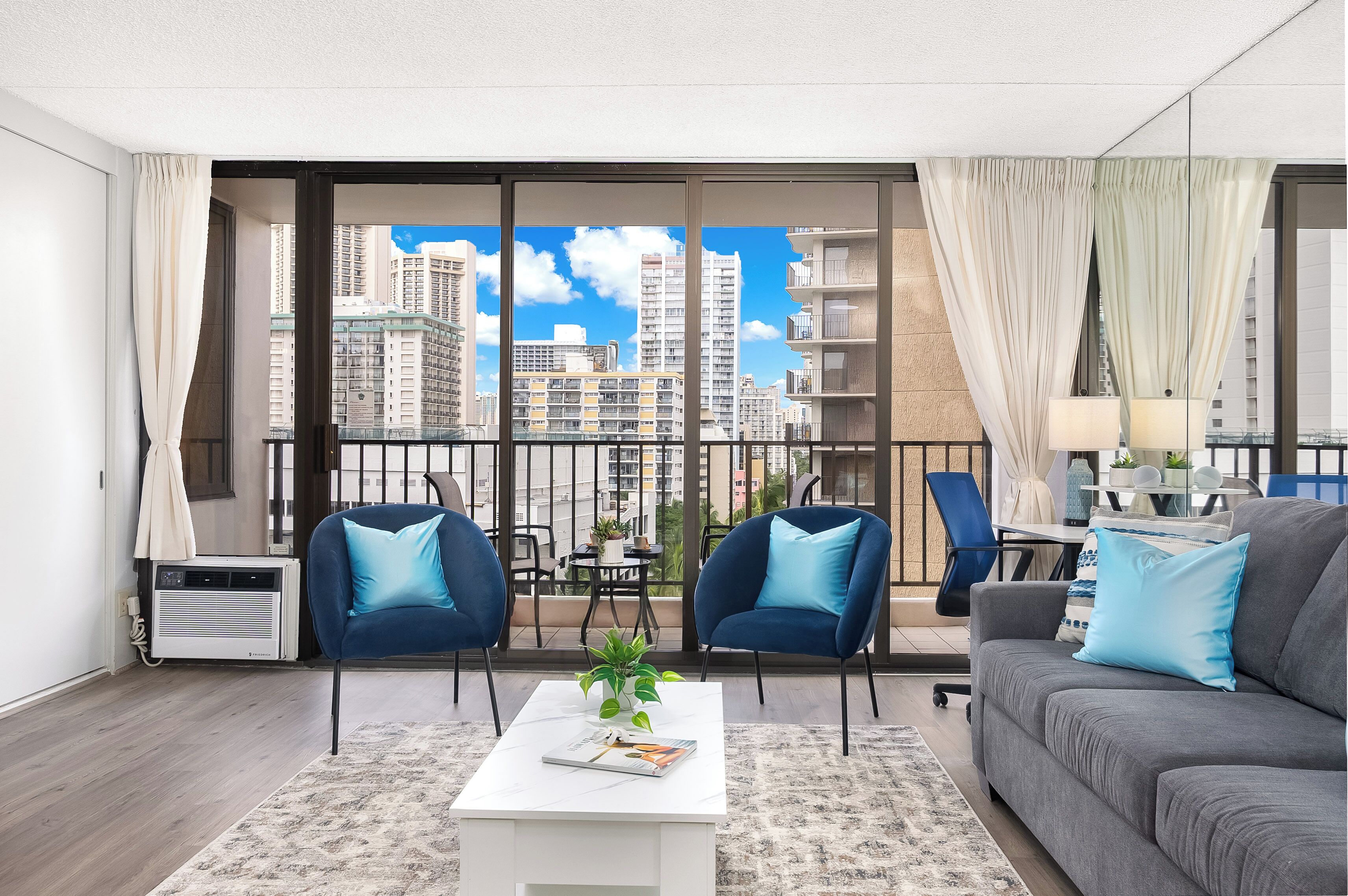 A modern living room with a grey sofa, two blue armchairs, a white coffee table, and a view of tall buildings through large windows.