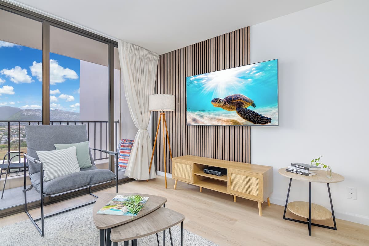 Un salon moderne avec un canapé gris, un meuble TV en bois, une petite table ronde et un balcon offrant une vue sur les montagnes.