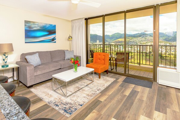 Una sala moderna con un sofá gris, una mesa de centro blanca y un sillón naranja. Un ventanal ofrece vistas a un paisaje urbano y montañas.