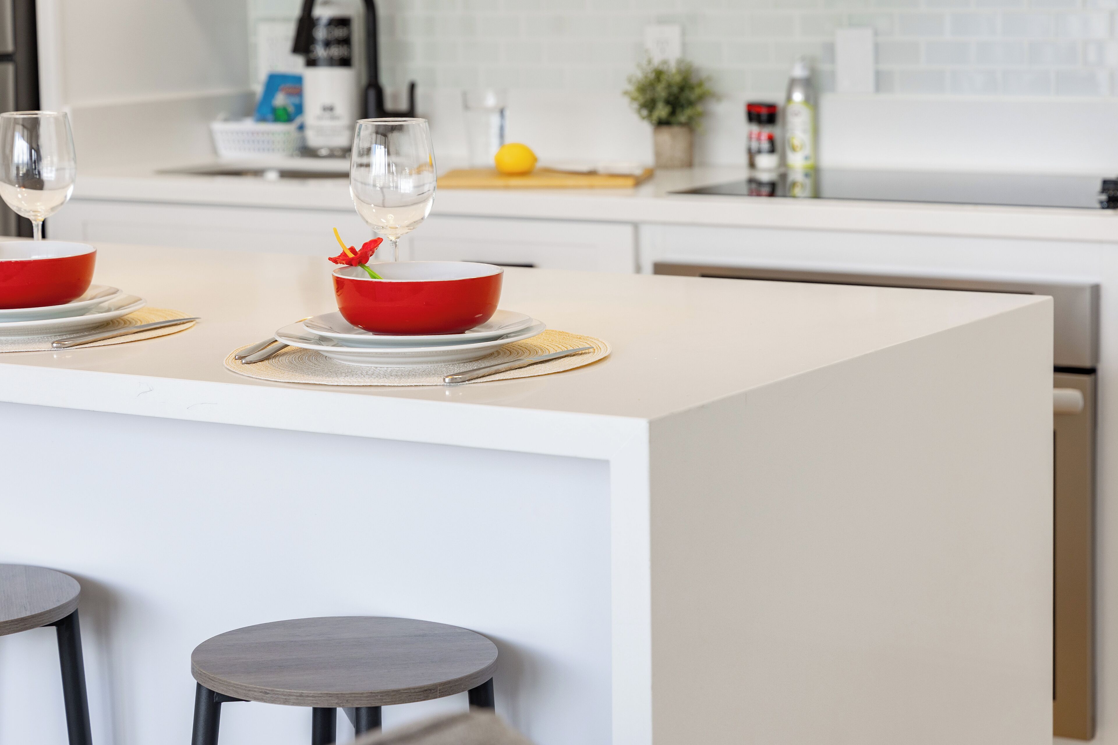 A modern kitchen with a central island, two bar stools, and a dining setup with red bowls and wine glasses.