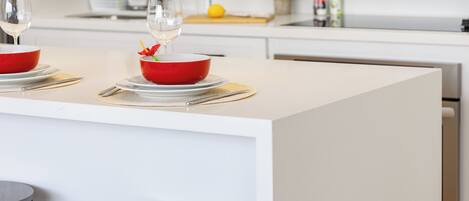 A modern kitchen with a central island, two bar stools, and a dining setup with red bowls and wine glasses.