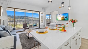 A modern living room with a large window, a sofa, a dining table with a fruit bowl and glasses, and a kitchen area with a TV. - High Floor Mountain View with King or Queen Bed and Parking (Honolulu)