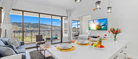 Una sala de estar moderna con un ventanal amplio, un sofá, una mesa de comedor con fruteros y vasos, y una zona de cocina con un televisor.