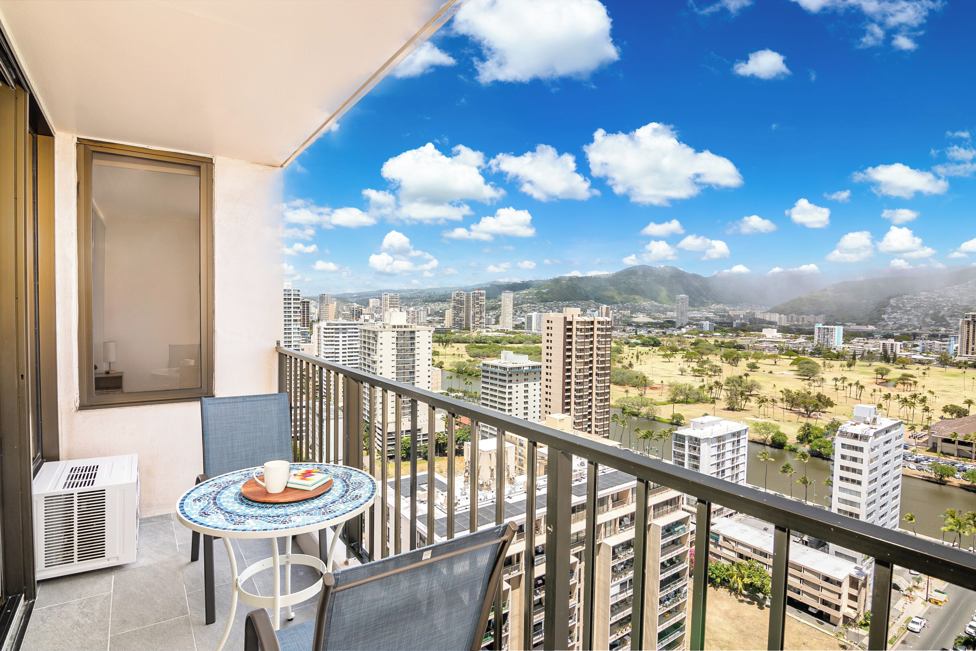 A balcony with a table and chairs, overlooking a cityscape with buildings and a river.