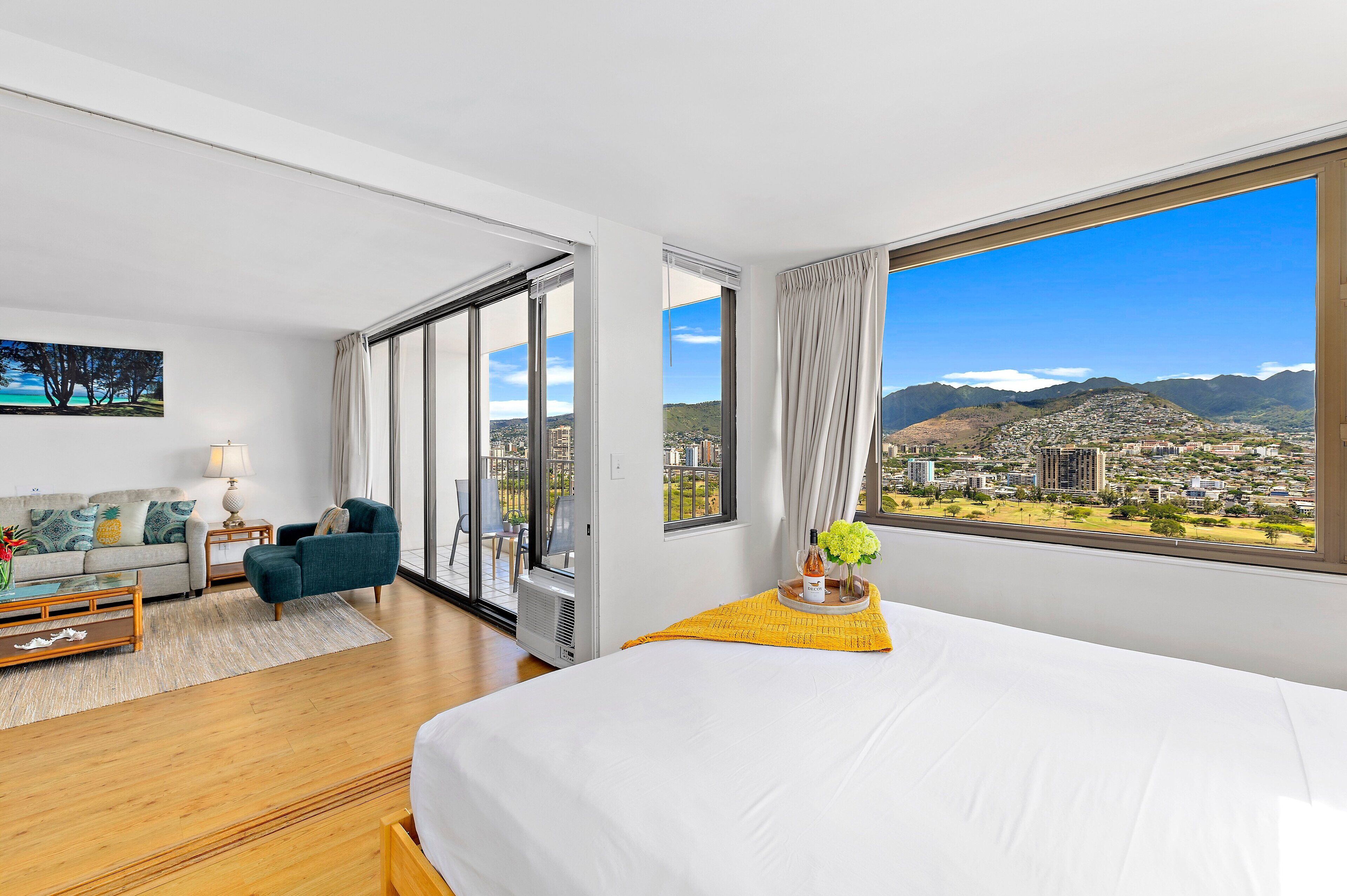 A hotel room with a large bed, a sofa, a coffee table, and a view of a cityscape with mountains.