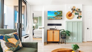 A modern living room with a green sofa, a dining table with fruit, a TV, and a wall-mounted guitar. - Mountain View Condo with King or Queen Bed and Parking (Honolulu)