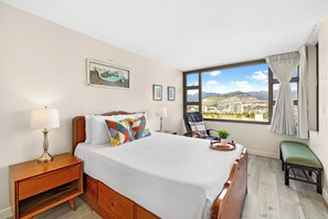 A hotel room with a large bed, a nightstand with a lamp, a chair, a bench, and a window with a city view. - Mountain View Condo with King or Queen Bed and Parking (Honolulu)