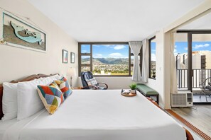 A hotel room with a large bed, a view of the city, a chair, a small table with a plant, and a window with curtains. - Mountain View Condo with King or Queen Bed and Parking (Honolulu)