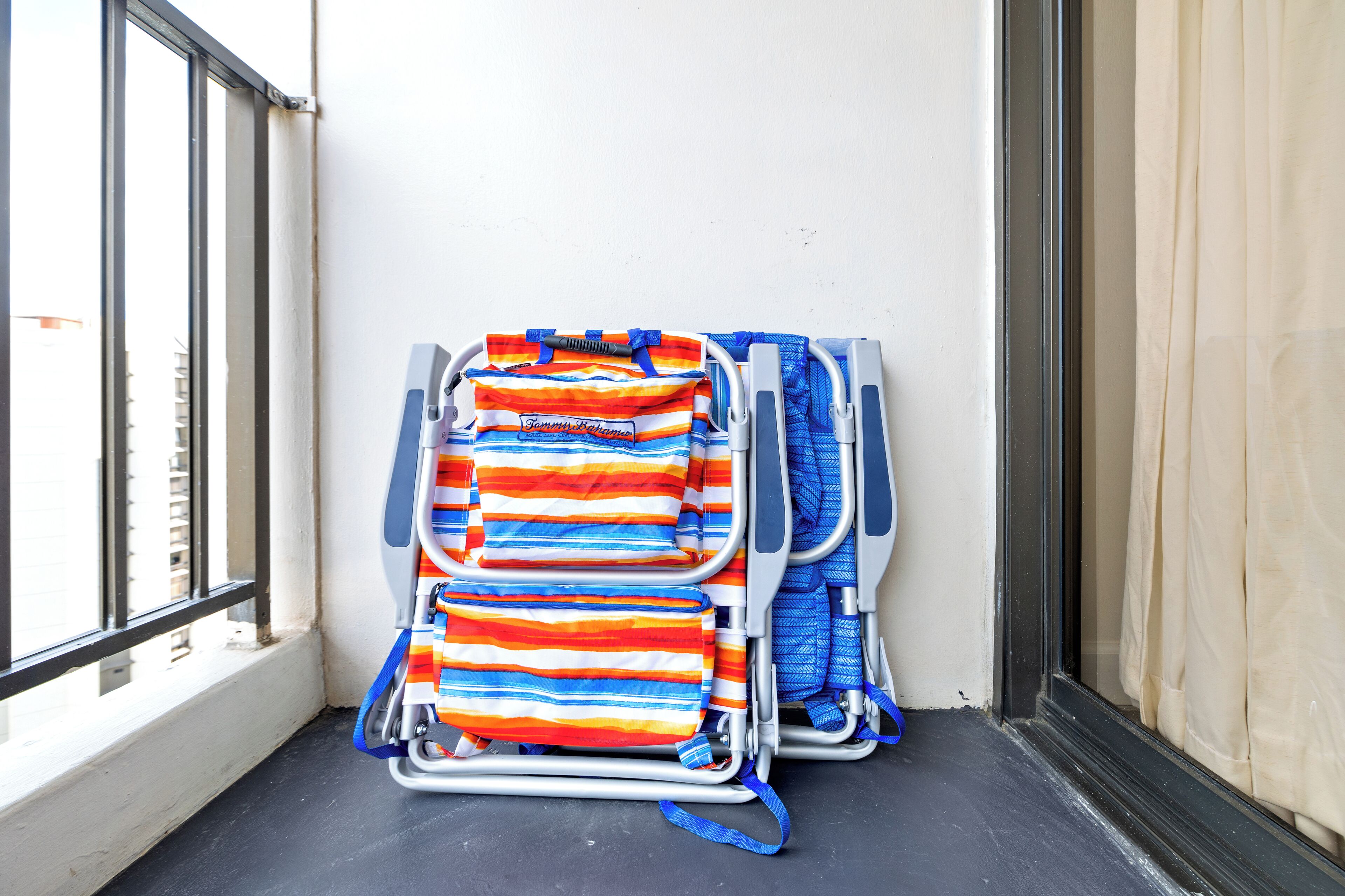 Un rincón de una habitación con una ventana, una puerta y un estante metálico con bolsas a rayas y una toalla azul.
