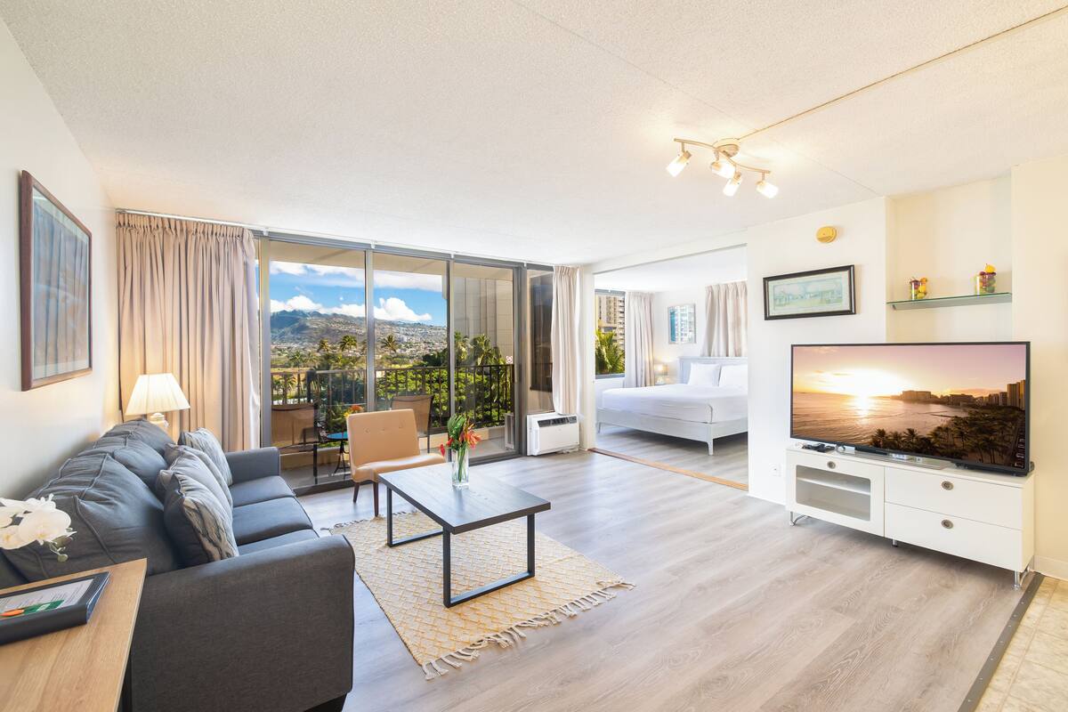 Ein modernes Wohnzimmer mit einer Couch, einem Couchtisch und einem Fernseher. Es gibt ein großes Fenster mit Blick auf die Stadt und einen Balkon.