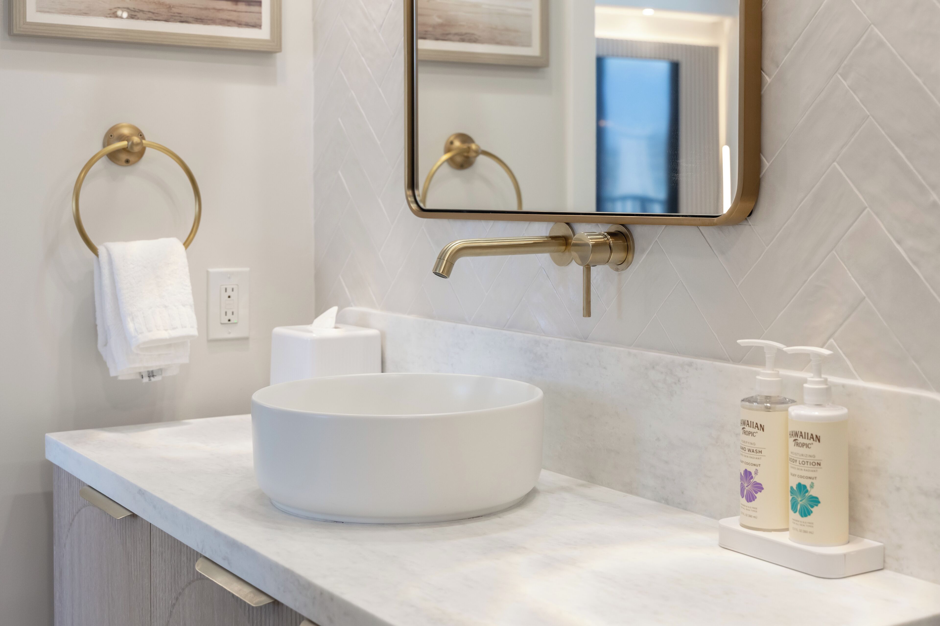 A bathroom with a white sink, gold towel ring, and two bottles of Hawaiian Tropic products on the counter.