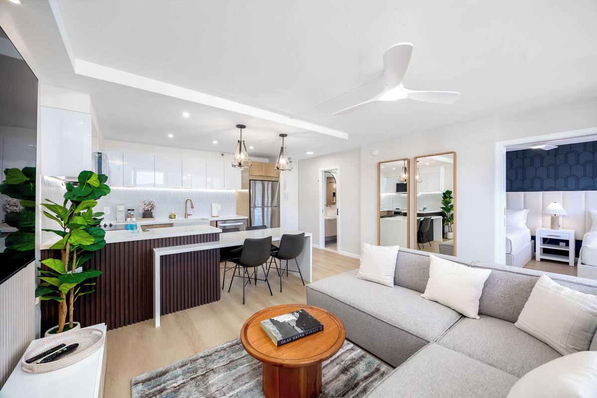 Ein modernes Wohnzimmer mit einer grauen Couch, einem Holztisch und einem Essbereich mit weißer Arbeitsplatte und schwarzen Stühlen.