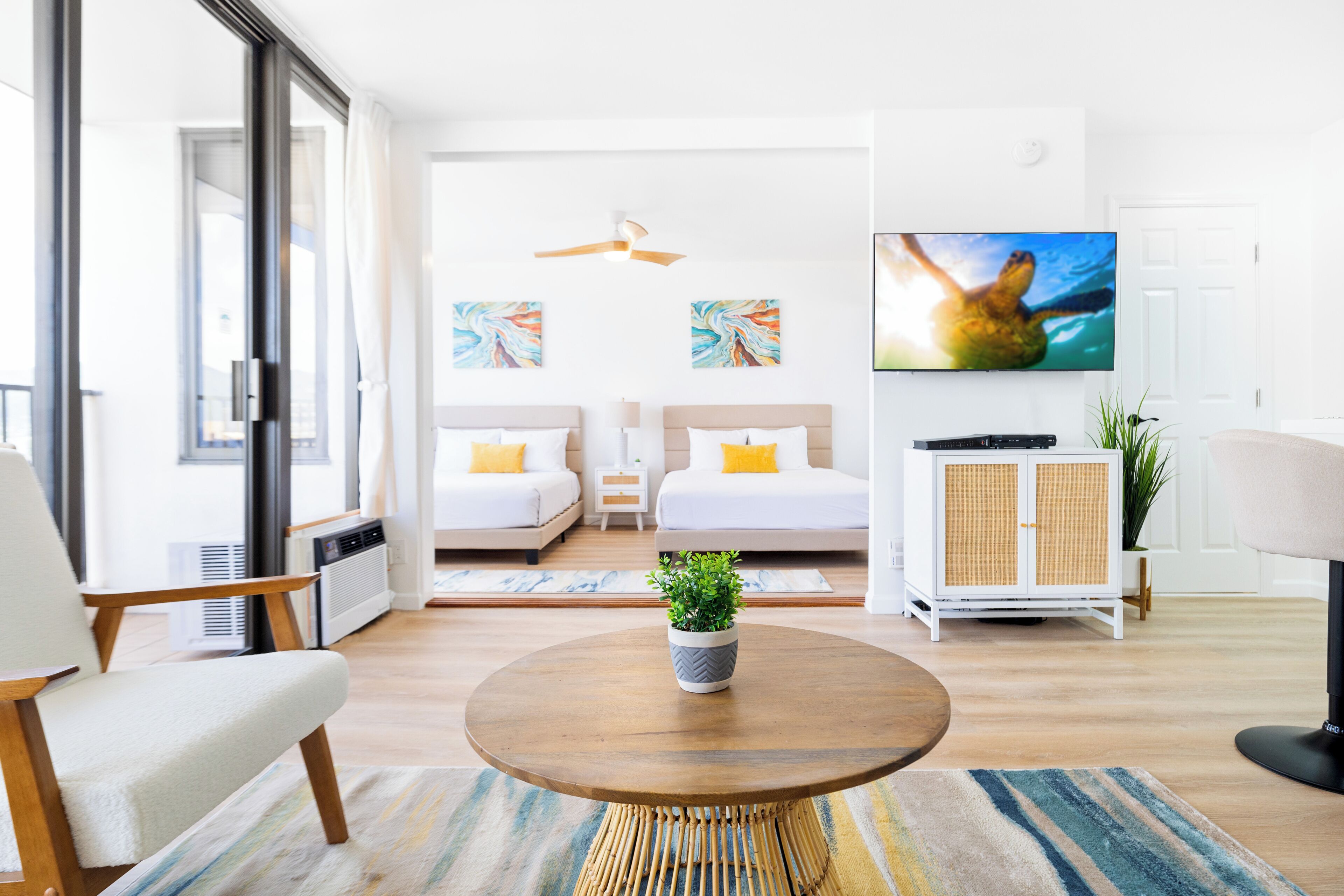 A modern hotel room with a large window, a flat-screen TV, two beds, a small cabinet, and a round coffee table.