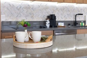 A kitchen with a modern design, featuring a herringbone-patterned backsplash, a coffee maker, and a wooden tray with two white mugs and a small plant. - Premium Remodeled Condo in Waikiki with 2 Beds and Parking (Honolulu)