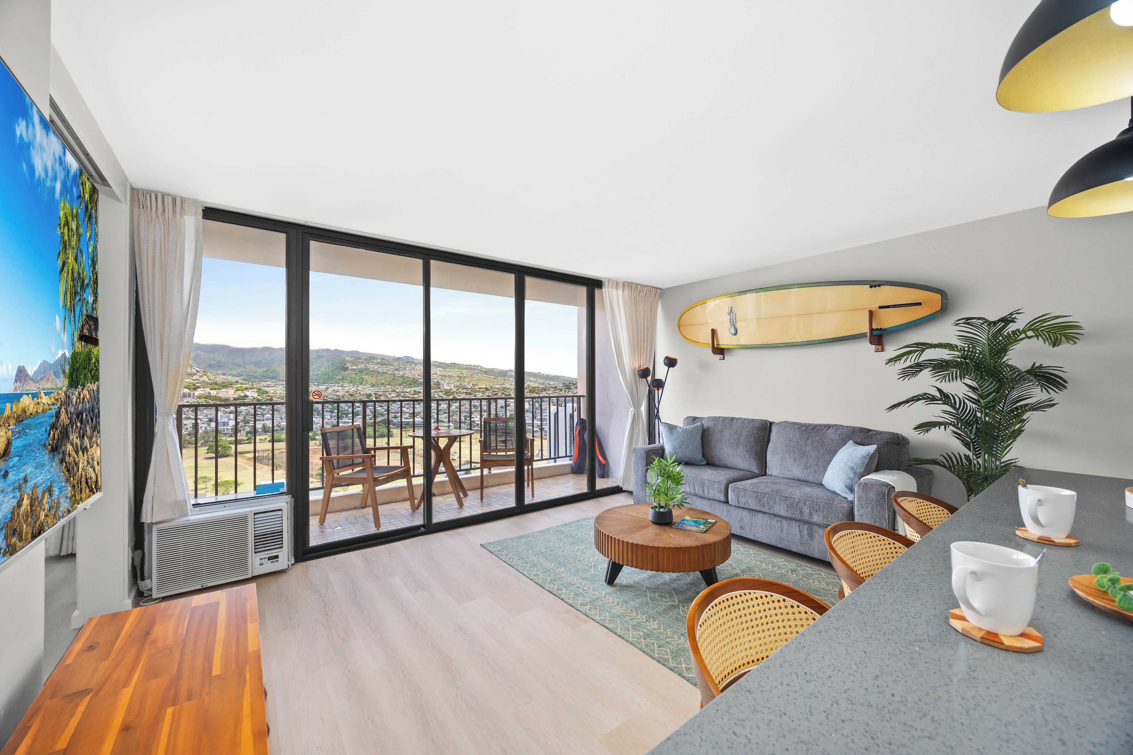 A modern living room with a balcony, a surfboard on the wall, and a view of the landscape.