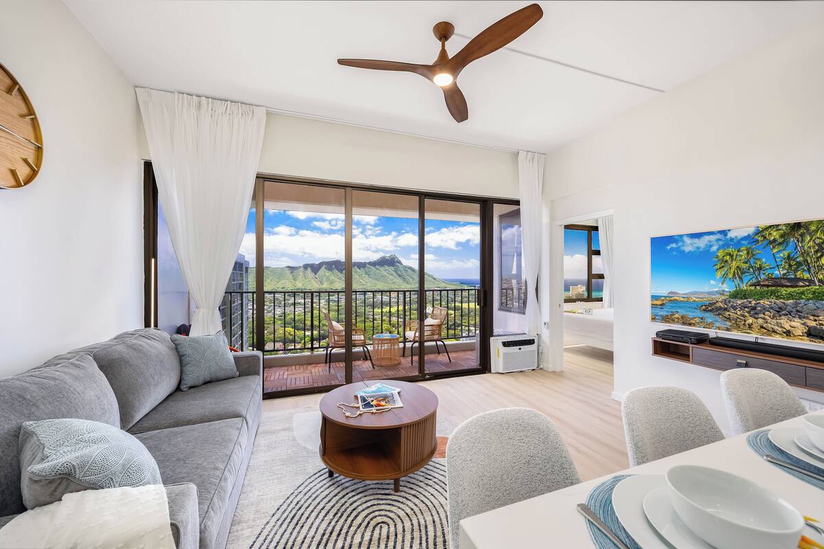 Ein modernes Wohnzimmer mit grauem Sofa, rundem Couchtisch und Essbereich mit weißen Tellern und Schüsseln. Ein Deckenventilator ist sichtbar, und das Zimmer bietet große Fenster mit Blick auf Berge und Grünflächen.