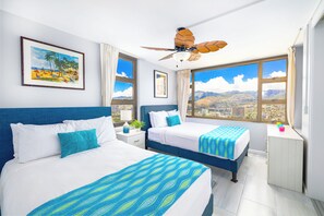 Ein Hotelzimmer mit zwei Betten, einem Deckenventilator, großen Fenstern mit Blick auf die Stadt und einem gerahmten Gemälde an der Wand.