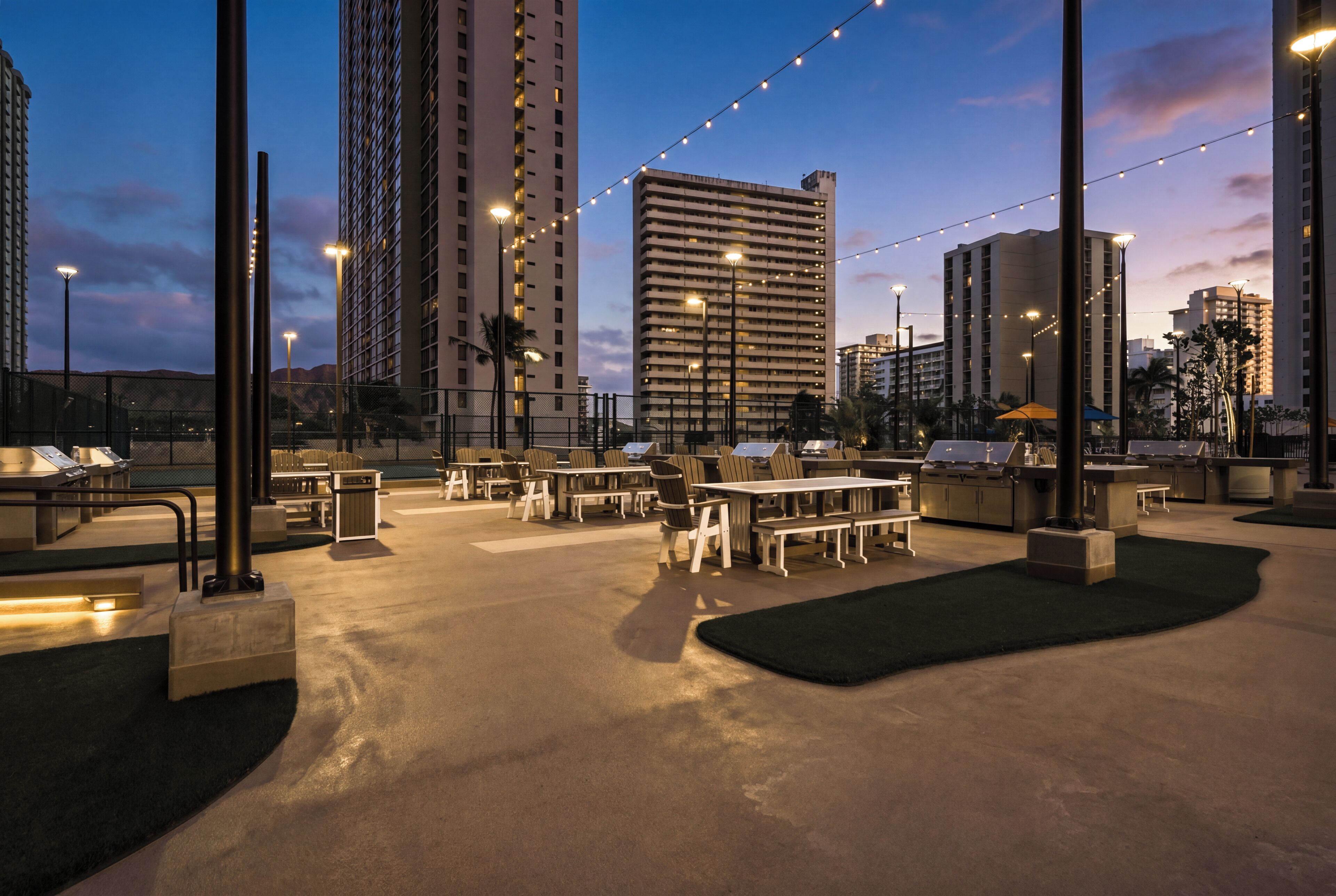 An outdoor dining area with tables and chairs, surrounded by tall buildings and string lights.