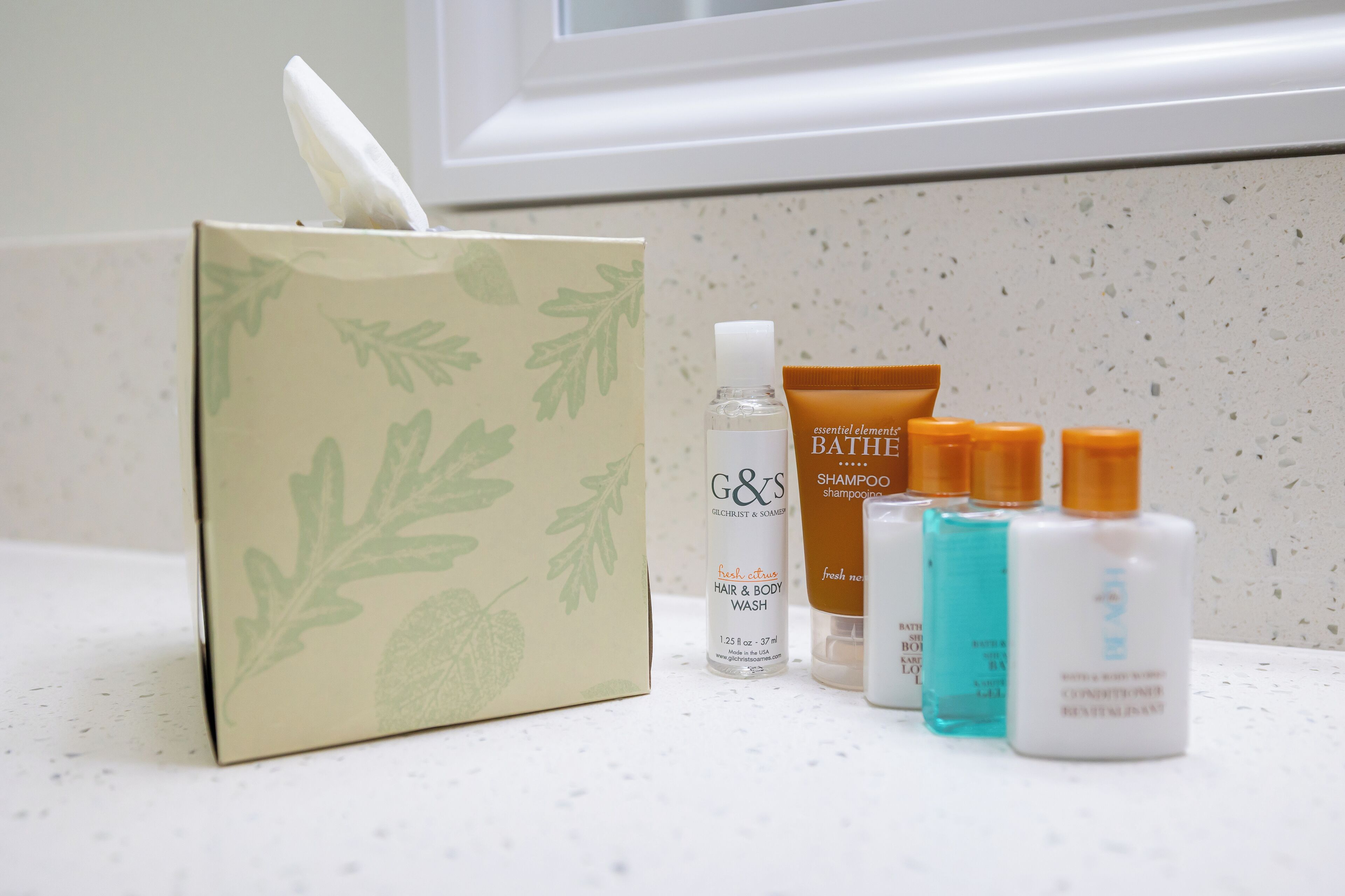 A bathroom counter with a tissue box, a bottle of G&J's fresh citrus hair and body wash, a Bath & Body Works shampoo bar, and a white bottle of lotion.