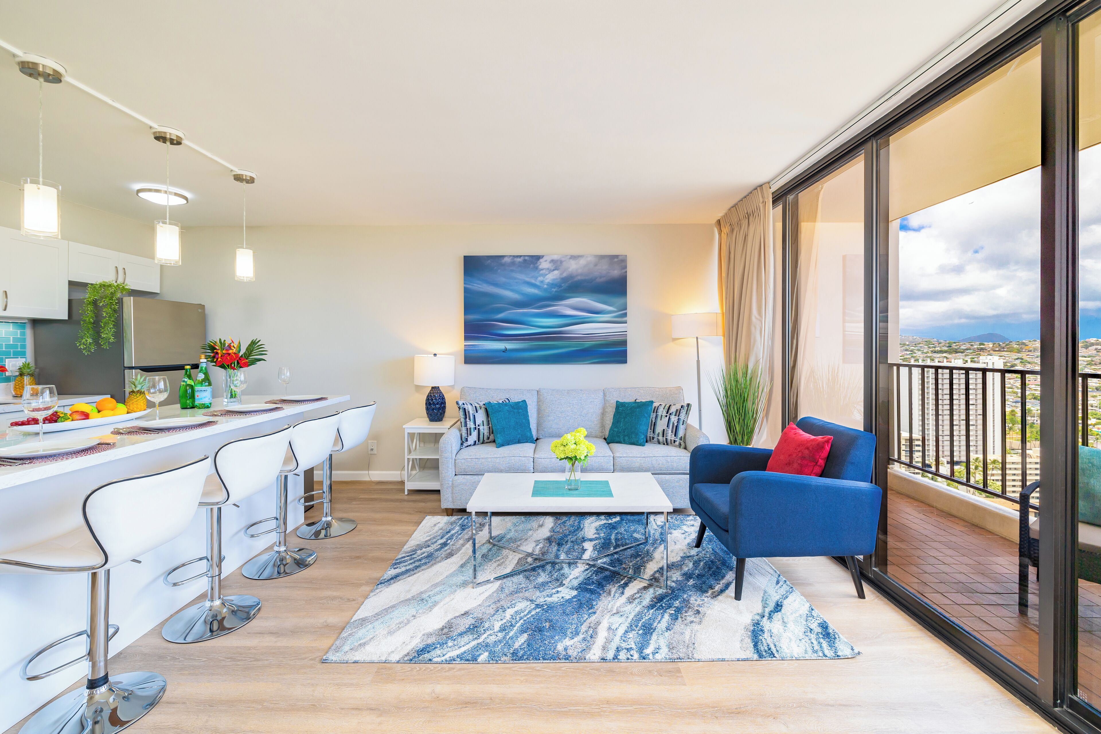 A modern living room with a kitchen area, a balcony with a view, and a large abstract painting on the wall.