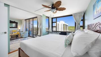 A hotel room with a bed, a desk, a chair, a ceiling fan, and a large window offering a view of buildings and greenery.