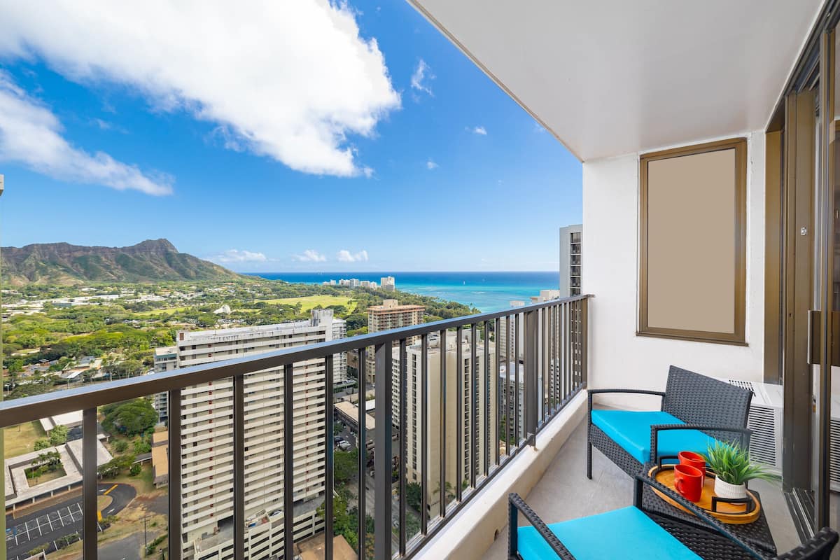 Uma varanda com vista para o oceano e montanhas, uma cadeira com almofada azul e uma pequena mesa com uma planta e duas chávenas vermelhas.