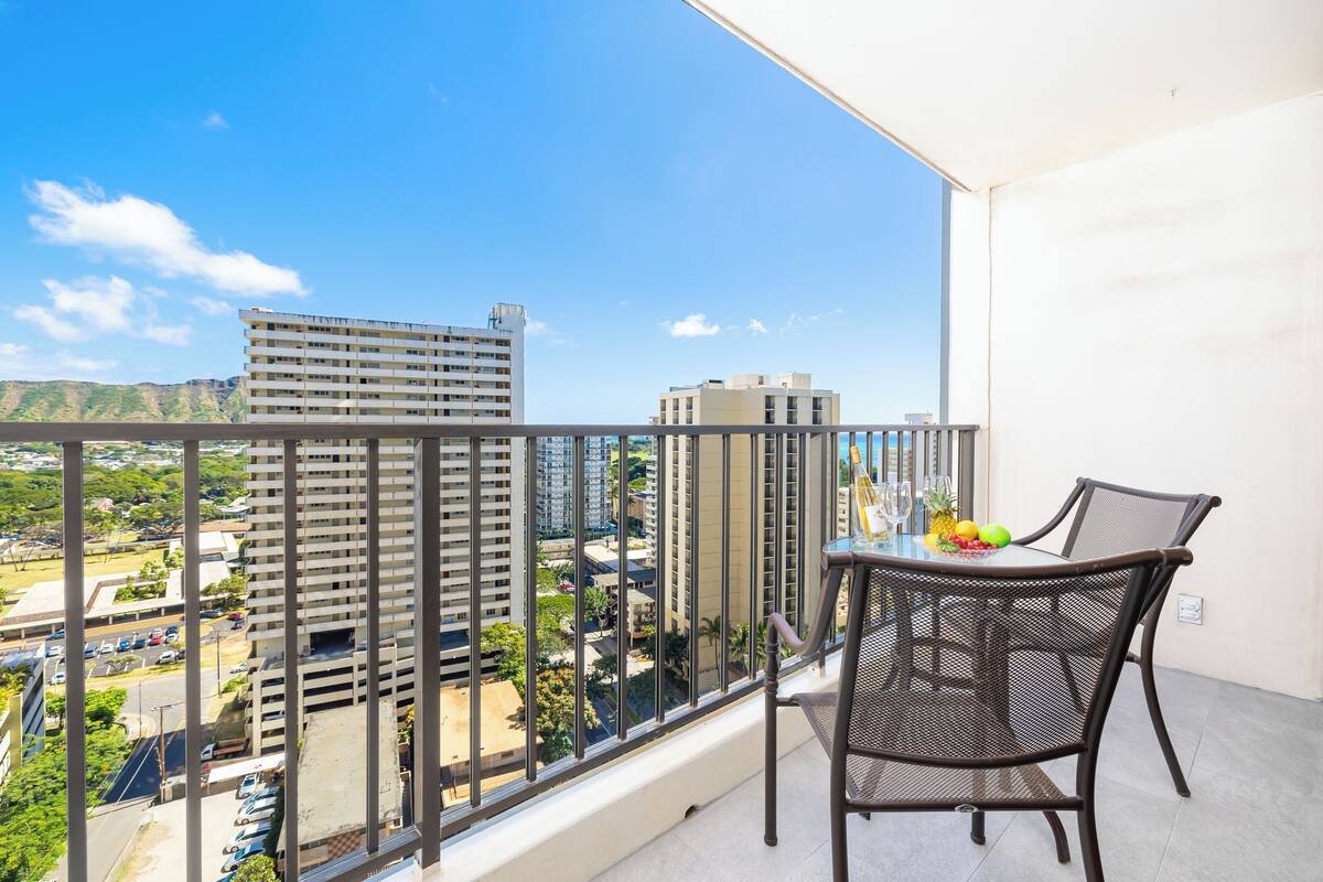 Un balcon avec une table et des chaises, donnant sur un paysage urbain avec de hauts immeubles et un ciel bleu dégagé.