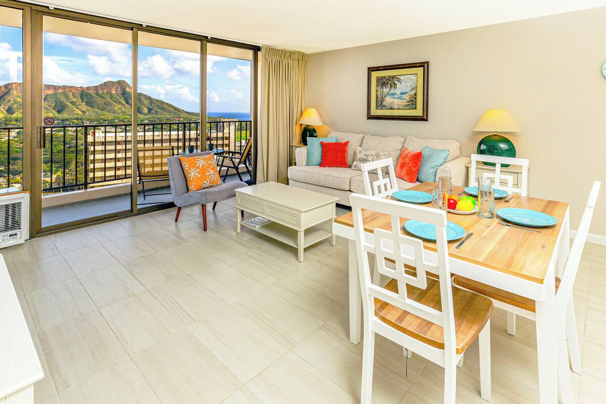 Un vaste espace de vie avec balcon, une table à manger pour deux personnes et un canapé agrémenté de coussins colorés.