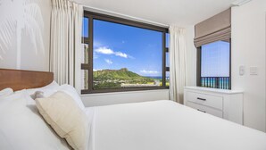 A hotel room with a large window, a bed, a nightstand, and a view of a mountain and beach. - Diamond Head and Partial Ocean views with 2 Beds and Parking (Honolulu)