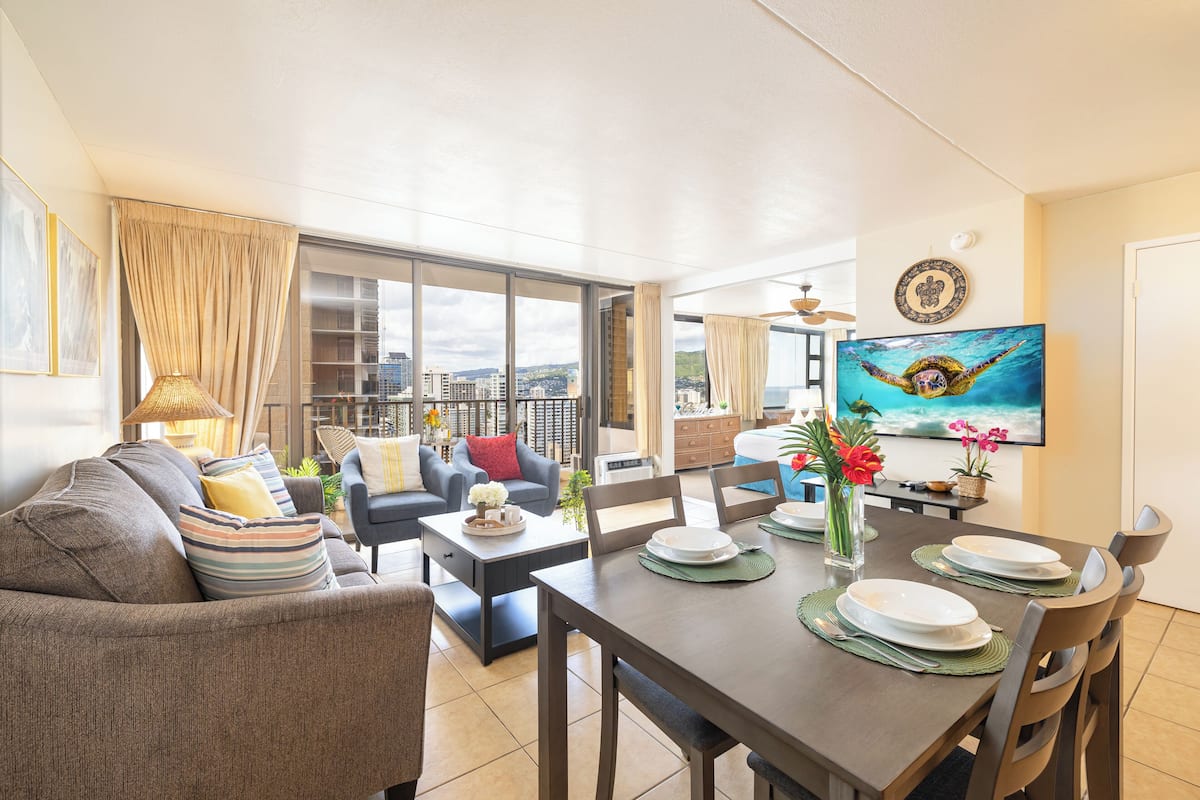 A spacious living area with a dining table set for four, a sofa, and a large window with a city view.