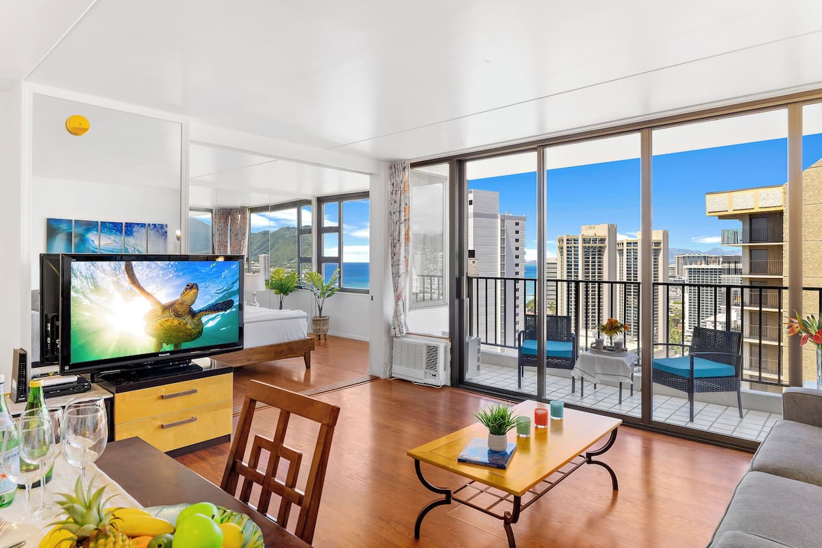 A modern living room with a large TV, wooden furniture, a balcony with a city view, and a dining area with a table and chairs.