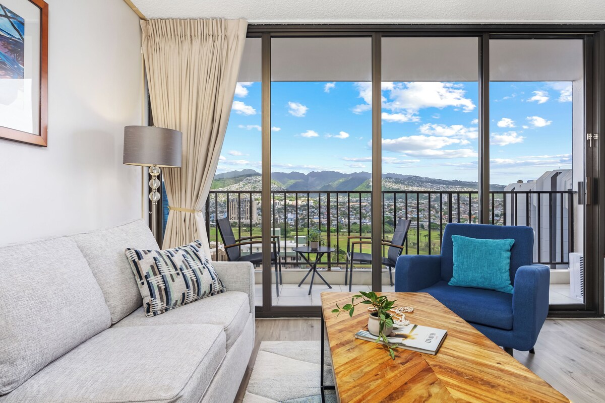 A modern living room with a sofa, a coffee table, and a balcony with a view of mountains and a cityscape.