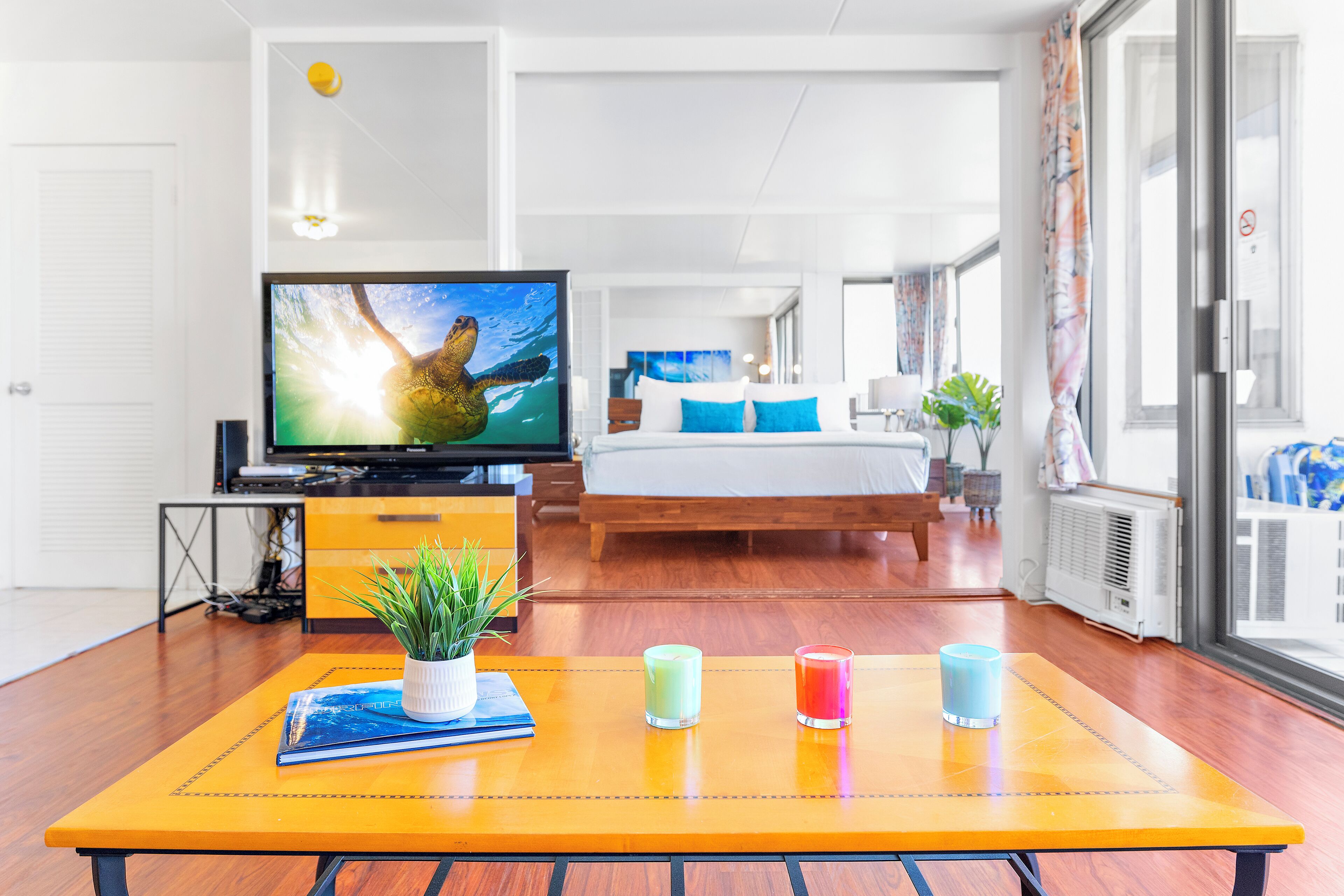 A modern hotel room with a wooden floor, a bed with blue and white bedding, a television on a stand, and a table with a plant and candles.