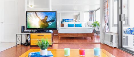 A modern hotel room with a wooden floor, a bed with blue and white bedding, a television on a stand, and a table with a plant and candles.