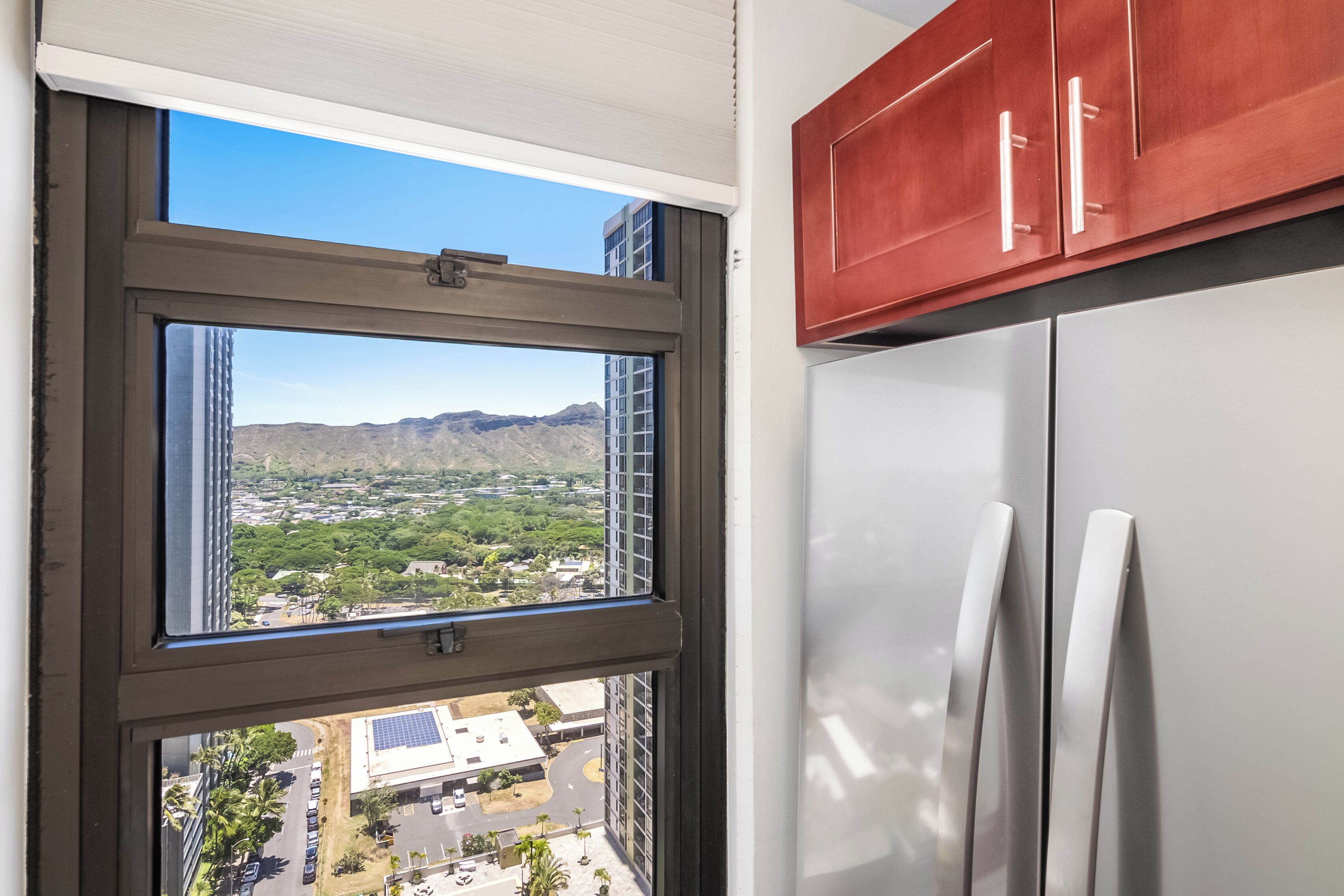 A kitchen with a view of a cityscape through a window, a refrigerator, and wooden cabinets.