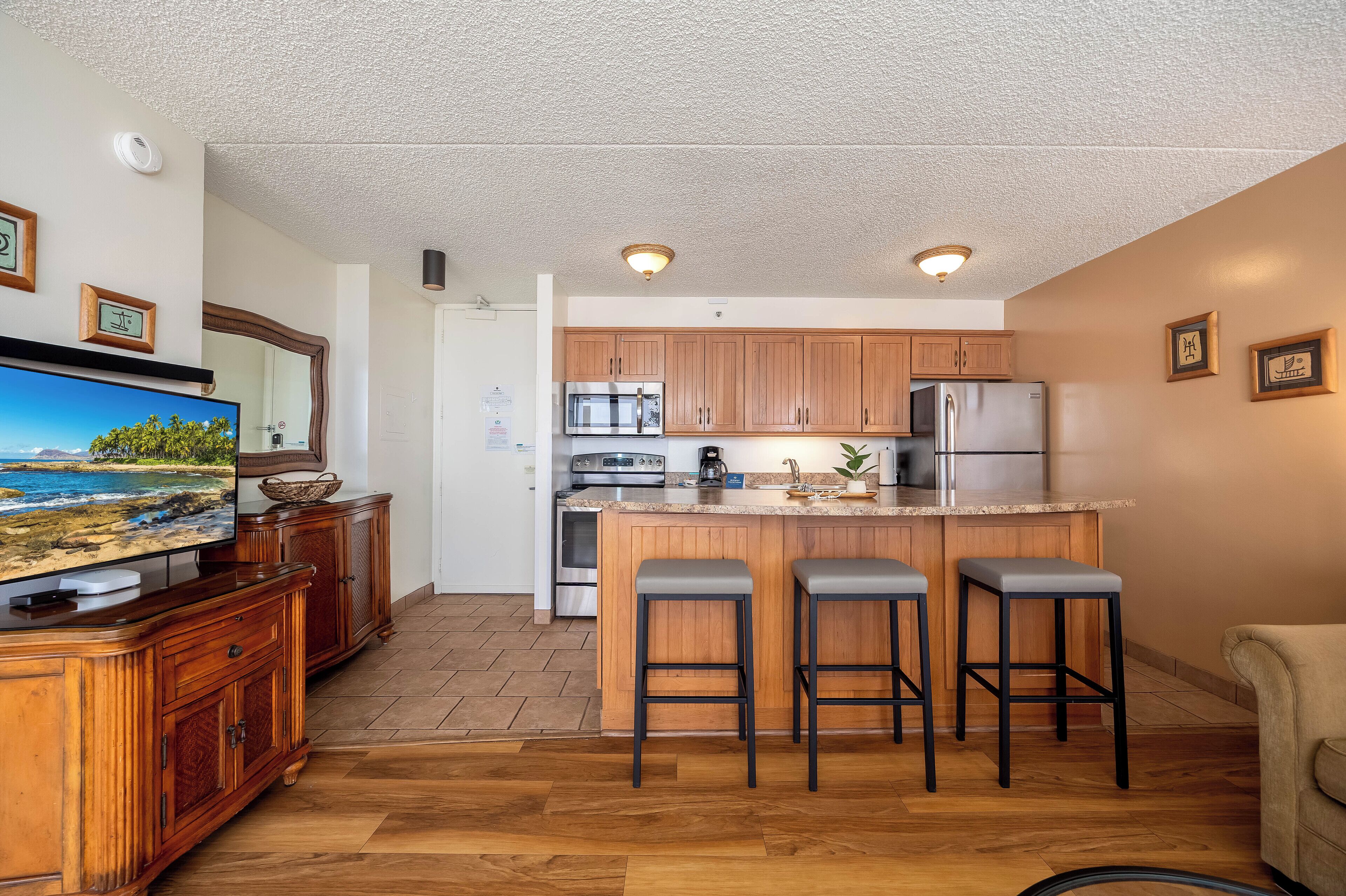 A modern kitchen with wooden cabinets, a built-in island with bar stools, a large flat-screen TV, and a ceiling-mounted light fixture.