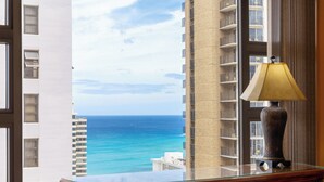 A view from a room with a large window overlooking the ocean, a lamp on a wooden desk, and a tall building in the background.