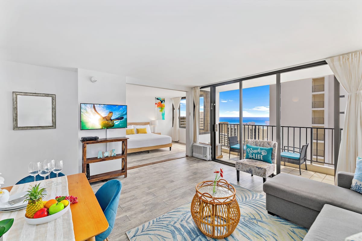 A modern hotel room with a balcony, a bed, a dining area with a fruit bowl, and a television.