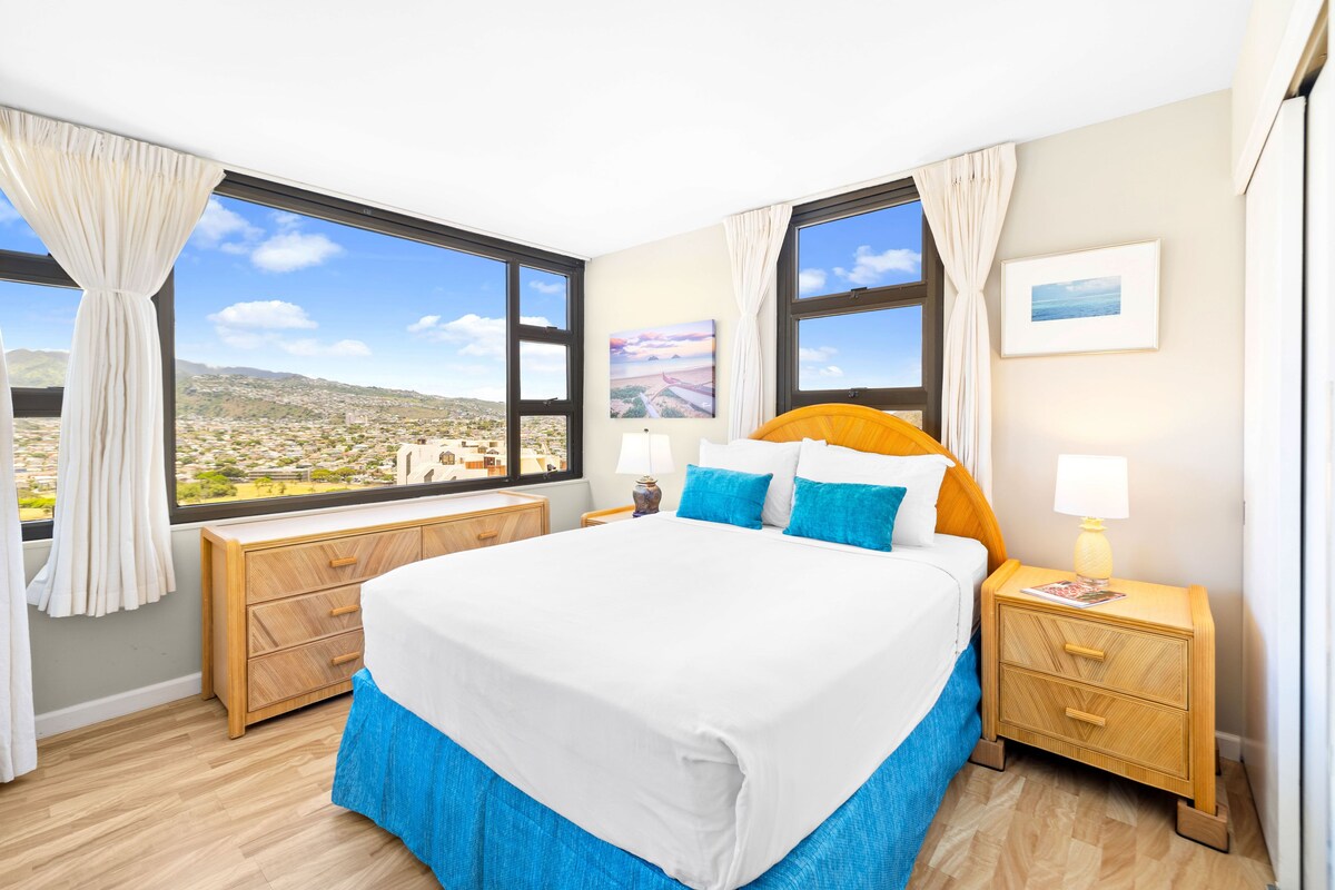 A hotel room with a large bed, two bedside tables, a view of a cityscape, and a window with curtains.