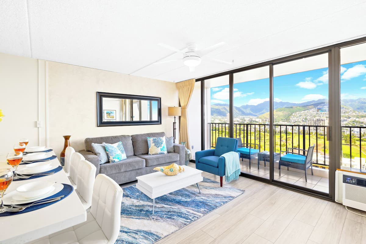 A modern living room with a dining area, a grey sofa, a blue armchair, and a balcony with a city view.