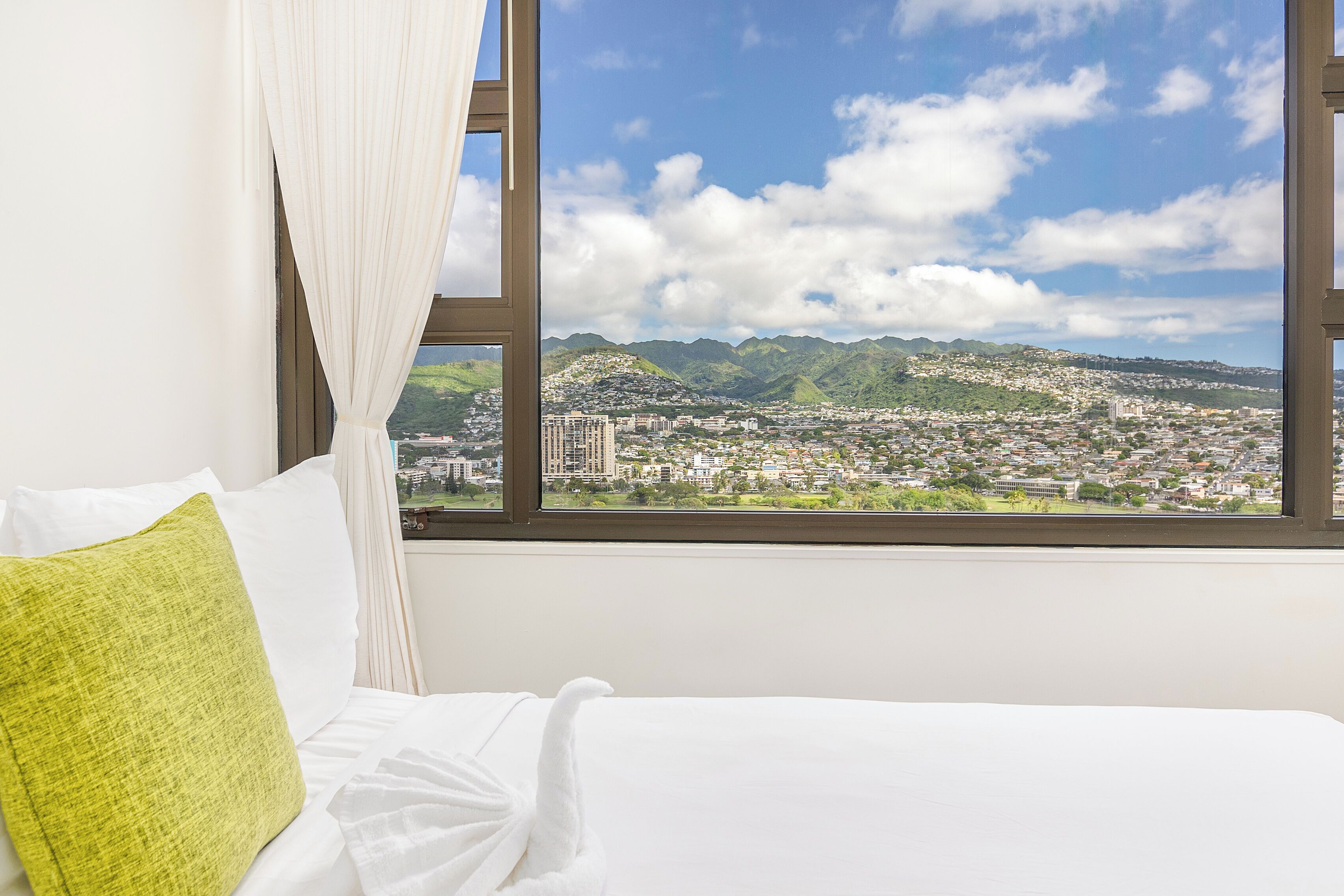A hotel room with a bed, a view of a cityscape through a window, and a curtain.