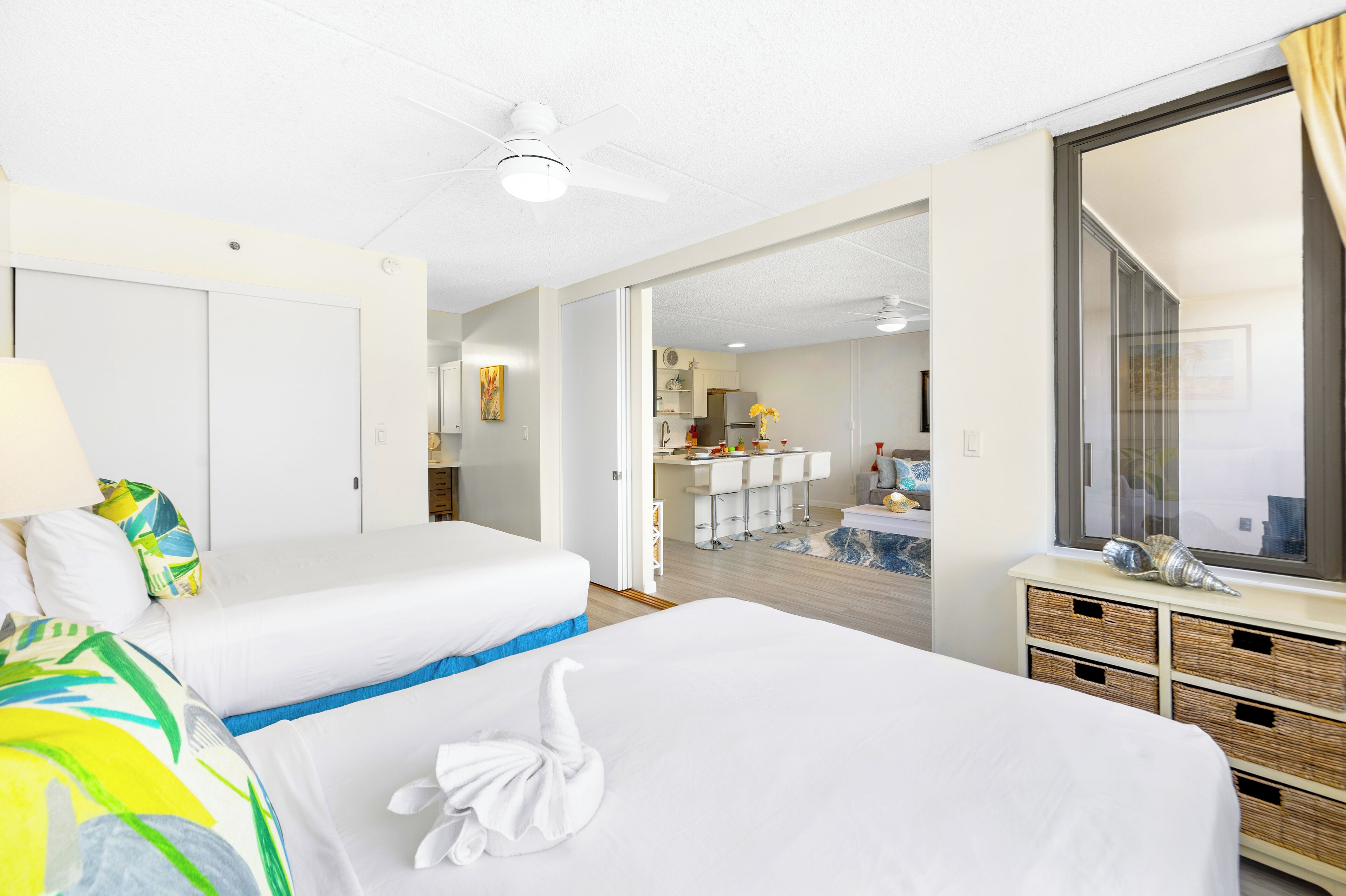 A hotel room with two beds, a desk, a kitchenette, and a large window with a view.