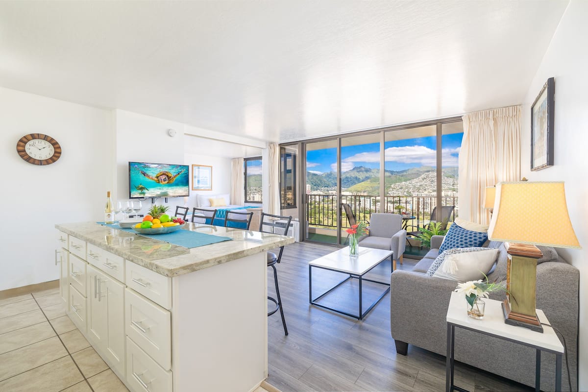 A modern living room with a kitchen area, a dining table, and a balcony with a city view.