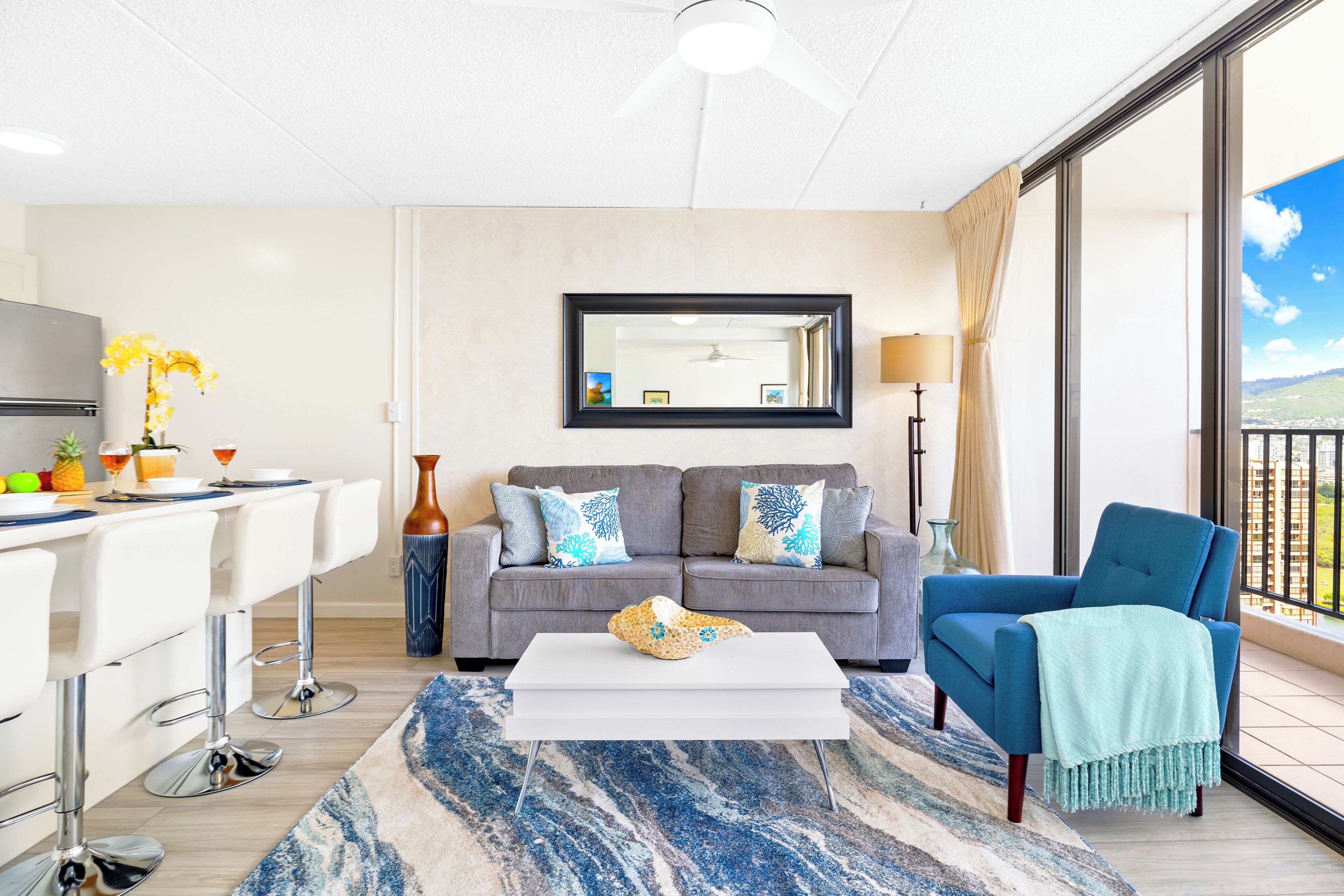 Een moderne woonkamer met een grijze bank, een witte salontafel en een blauwe fauteuil. Er is een groot raam met uitzicht op de omgeving.