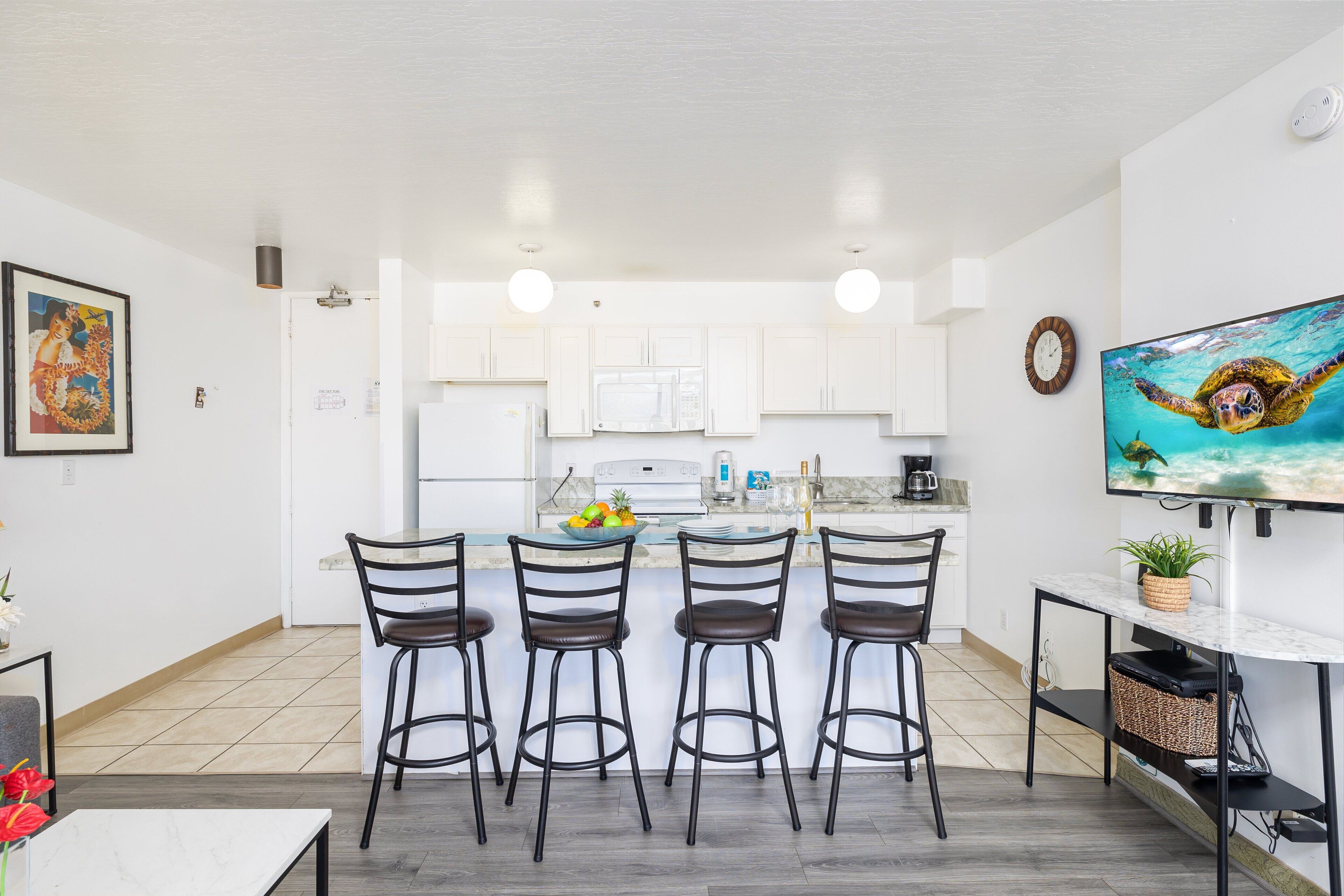 A modern kitchen with white cabinets, a central island with bar stools, a flat-screen TV, and a painting on the wall.