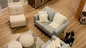 Een moderne woonkamer met een grijze zithoek, een witte fauteuil, en een keuken met een koelkast, oven en kasten.