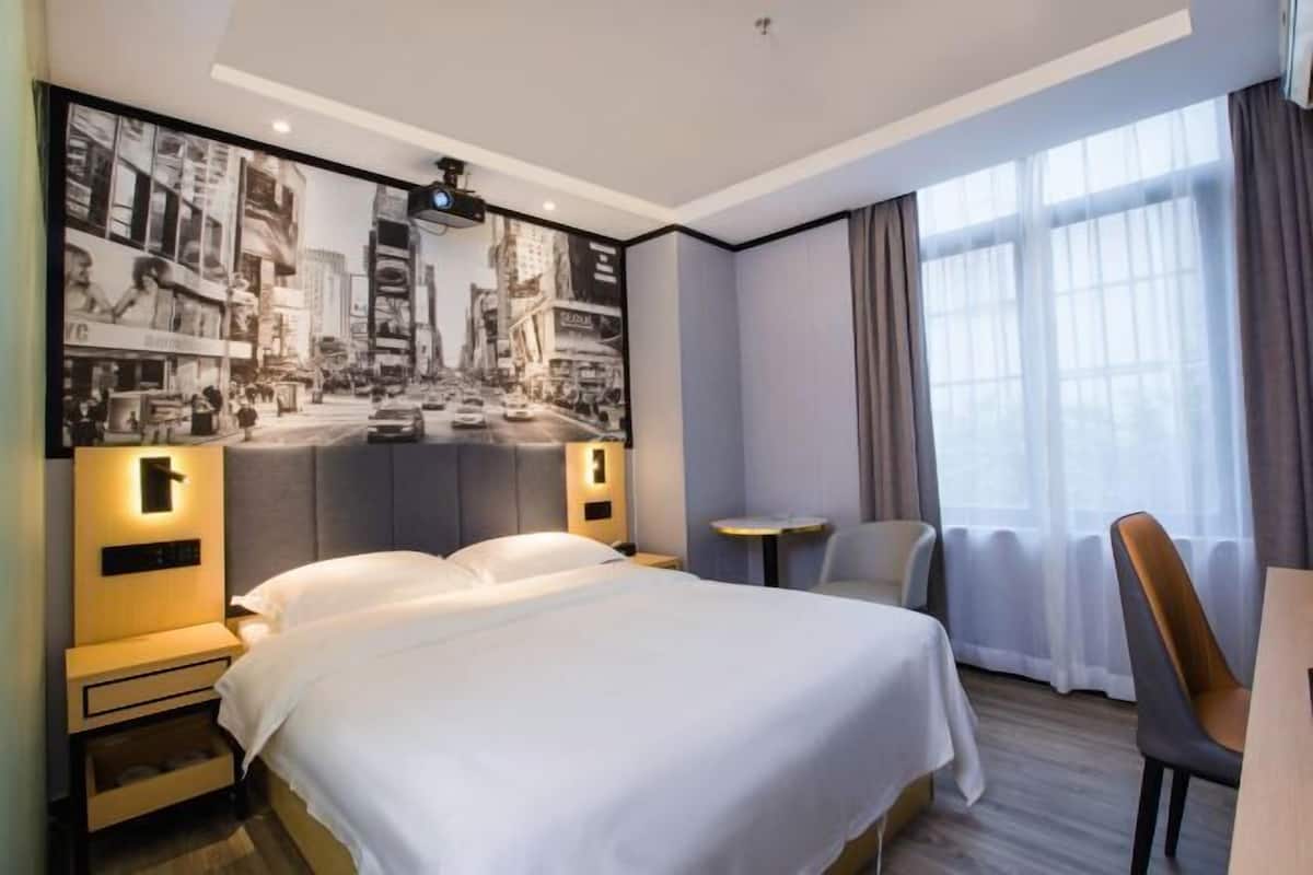 A hotel room with a large bed, a desk, a chair, a small table, and a large window with curtains.